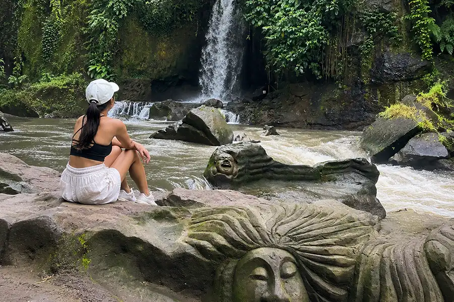 A woman in a white cap sitting on river rocks carved with faces, looking towards a waterfall pouring into a rushing river.