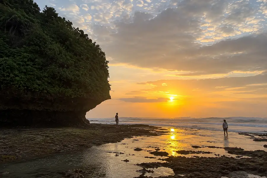 Silhouettes of visitors standing on a rocky beach reef during a brilliant golden sunset near a cliff.