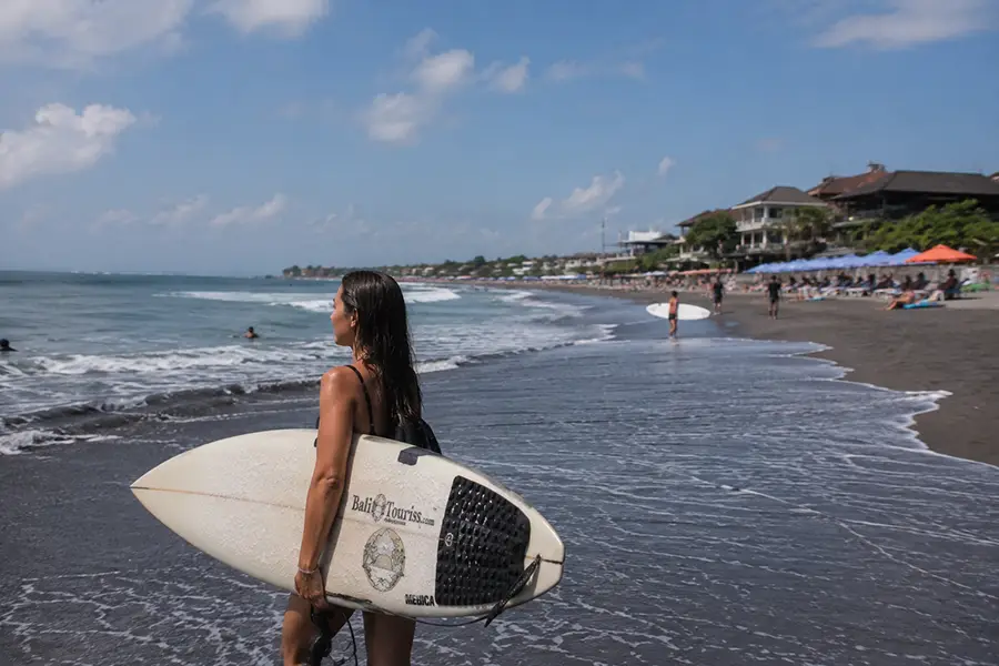 A woman holding a surfboard while standing on a volcanic sand beach looking at the ocean.