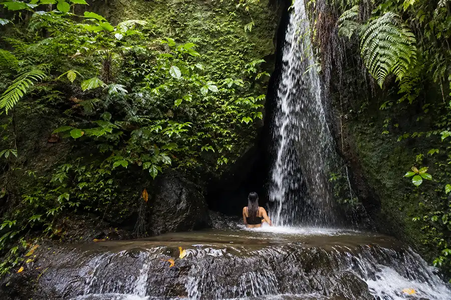 A woman sitting in a natural rock pool right under a narrow waterfall surrounded by lush green foliage.