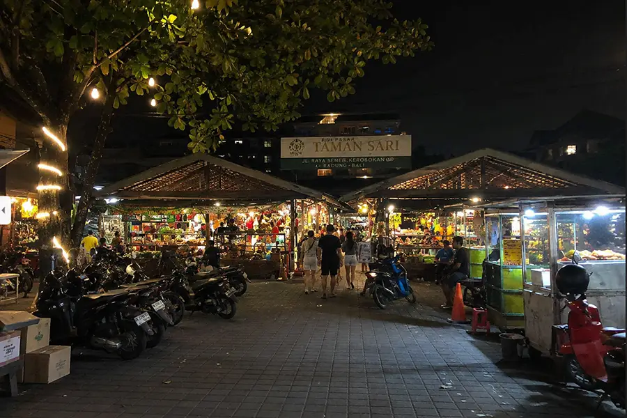 Pedestrians walking toward wooden market pavilions flanked by parked scooters and large shade trees.