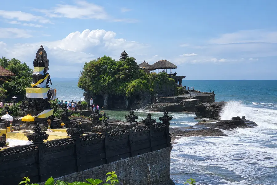 Ocean waves crashing against a rocky offshore temple under a blue sky, with a yellow-and-white draped shrine in the foreground.