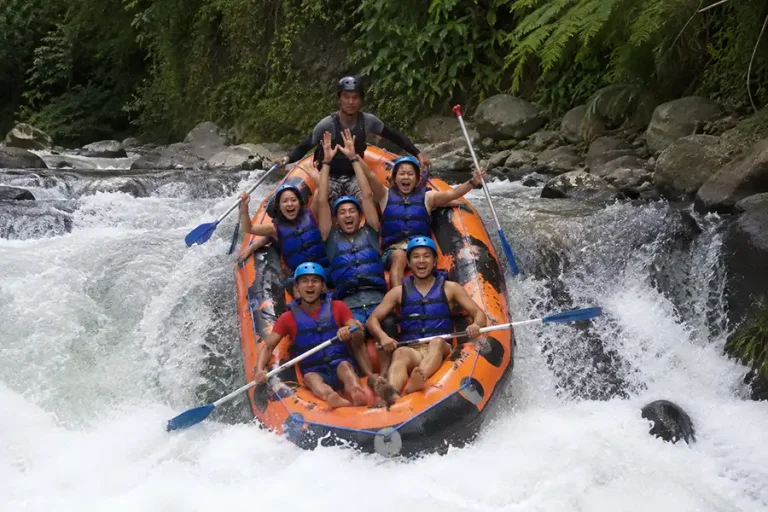 A team of rafters in blue safety gear shouting with joy as their orange raft splashes down a small river waterfall.