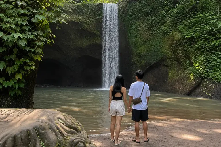 A couple holding hands on a sandy shore looking at a straight curtain waterfall flowing into a pool.