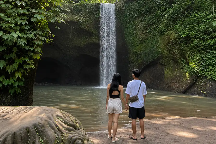 A couple holding hands on a sandy shore looking at a straight curtain waterfall flowing into a pool.