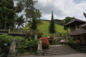 Two women in traditional Balinese clothing carrying ceremonial offerings on their heads past stone guardian statues.