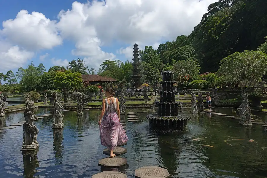 A woman in a long patterned pink dress walking across polygonal stone steps over a large pond with tier fountains.