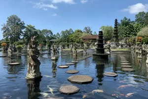 An ornate stone fountain and matching statues standing in a clear pond filled with large koi fish and concrete stepping stones.