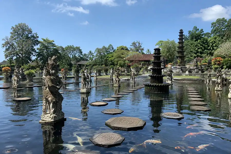 An ornate stone fountain and matching statues standing in a clear pond filled with large koi fish and concrete stepping stones.