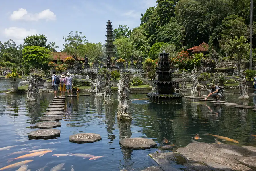 Several tourists standing and crouching on stepping stones in a garden pond to watch massive orange and white koi fish swim below.