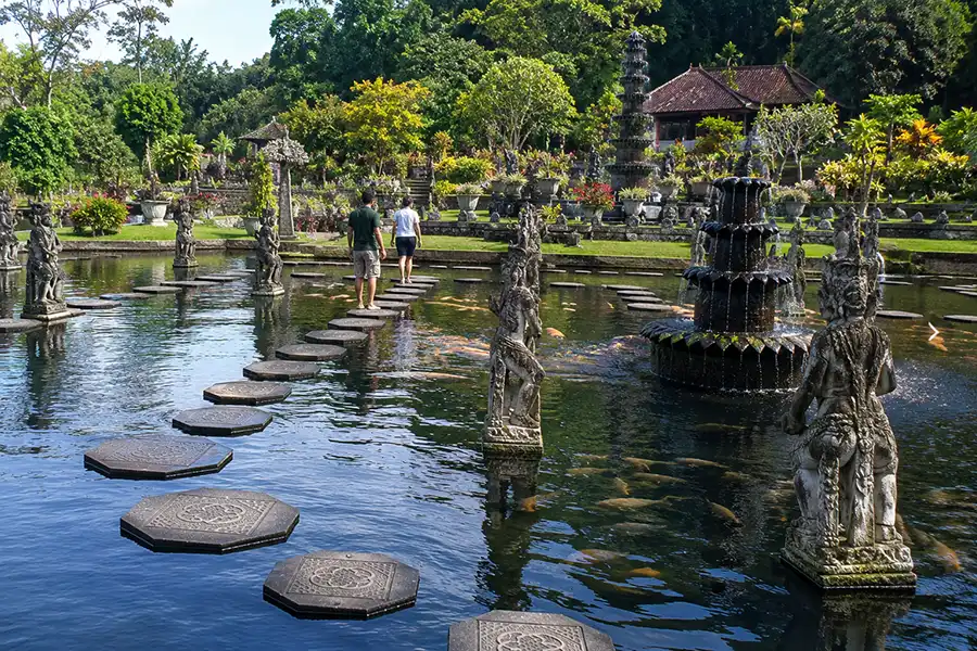 Visitors walking along a pathway of stone stepping stones over a large koi pond at Tirta Gangga during cultural tours in bali.