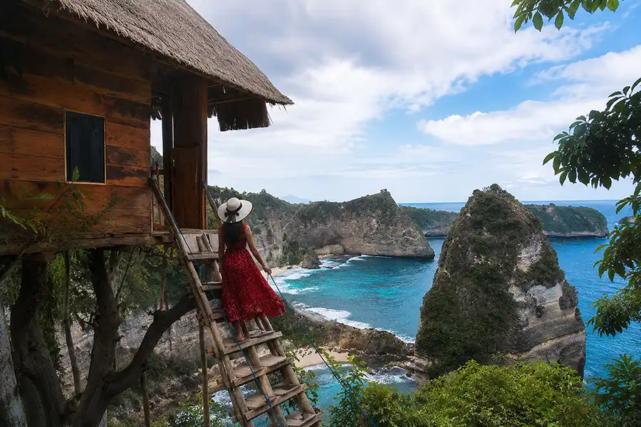 A woman in a red dress and white sun hat standing on the wooden stairs of a treehouse, looking out over coastal cliffs and blue ocean.