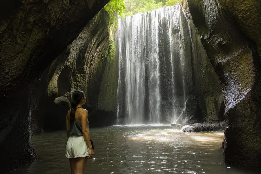 A woman from behind standing inside a dark rock cavern looking at a large tukad cepung waterfall.