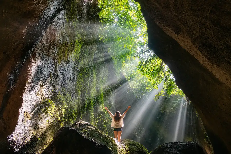 A woman standing on a mossy boulder inside a dark cave canyon with her arms raised, bathed in dramatic, bright sun rays.