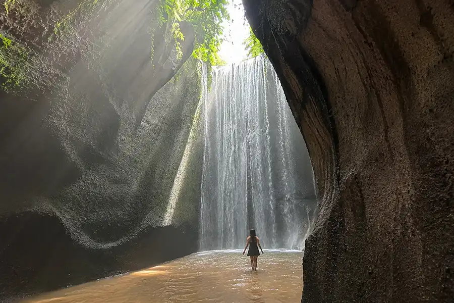 A woman standing in a shallow river inside a cave with sunbeams shining down on a wide waterfall.