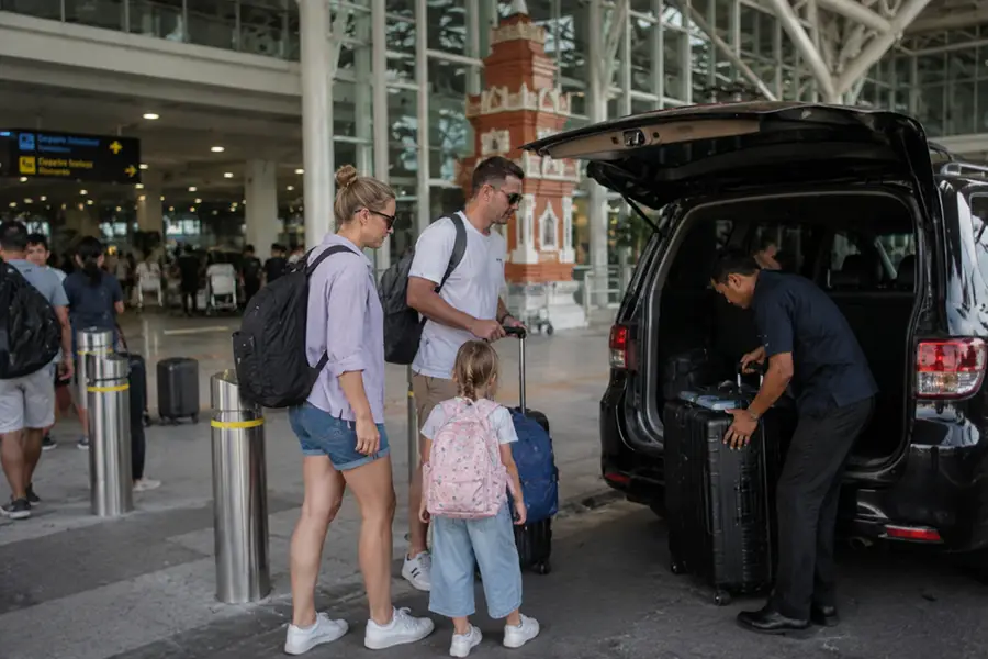 A helpful local driver loading heavy suitcases into the trunk of an SUV while a family with a young girl waits for their bali airport transfer with baby seat.