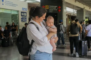 A mother holding her infant close while walking through the busy domestic arrivals terminal, looking for her pre-booked bali airport transfer with baby seat.