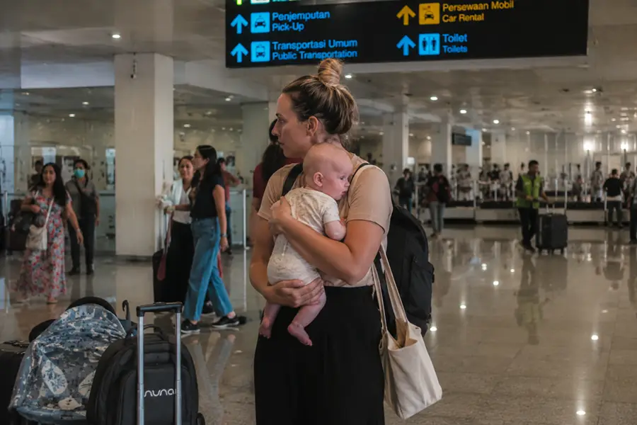 A mother cradling her baby inside the spacious terminal building near directional signs, ready to meet her driver for a bali airport transfer with baby seat.