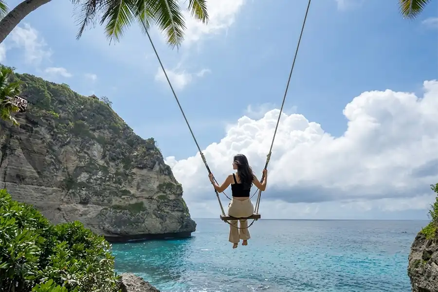 A woman on a high rope swing looking out at a bright blue ocean and sky.