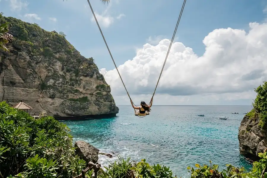 A woman soaring on a giant cliffside swing over a turquoise tropical bay with boats.