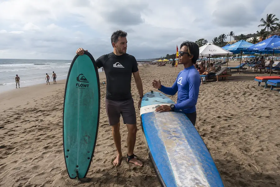 A male tourist and his surf instructor holding their boards during a beach lesson.