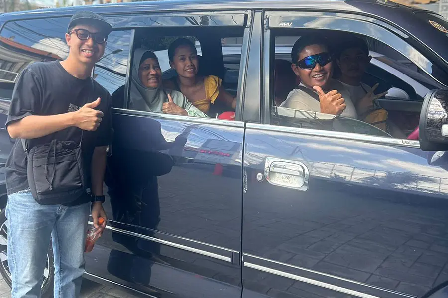 A group of smiling tourists and their driver giving thumbs-up signs from inside and next to a dark blue minivan.