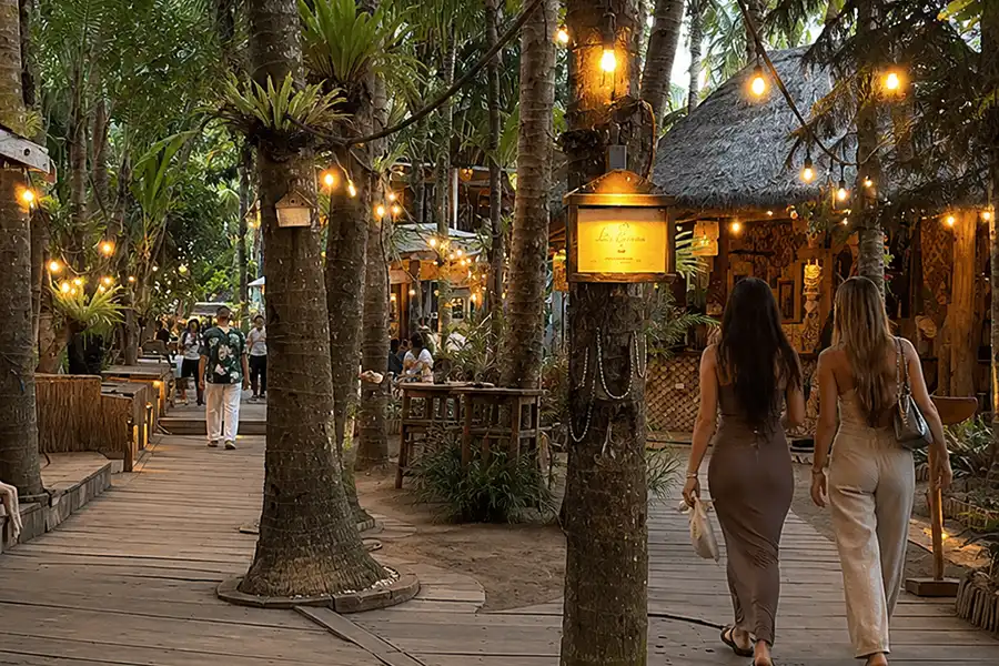 Two women walking down a wooden boardwalk inside an eco-style beach club decorated with hanging fairy lights and palm trees.