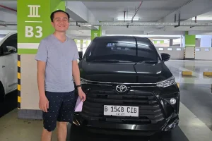 A man in a grey t-shirt standing next to a sleek black MPV inside an indoor airport parking garage.