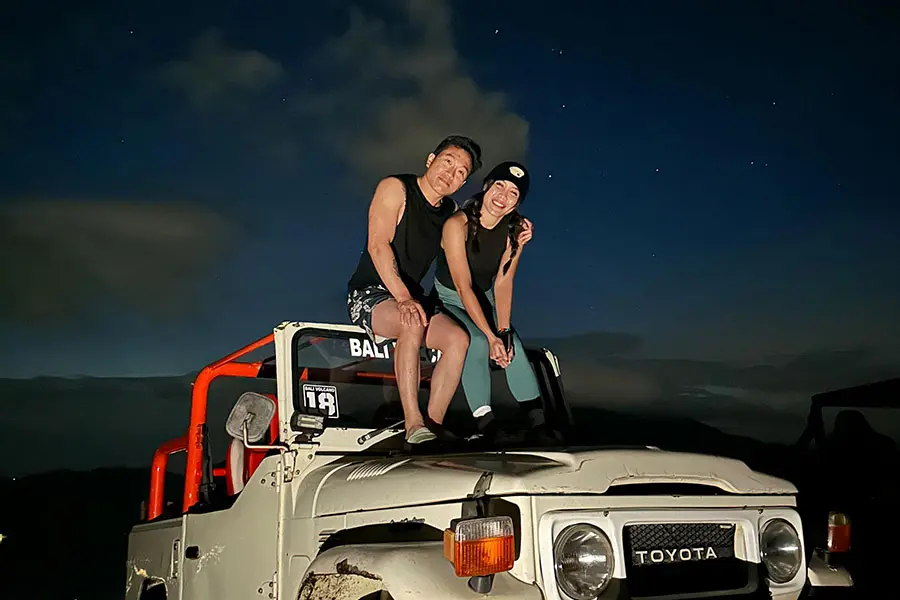 A smiling couple sitting on top of a classic white 4x4 Jeep under a dark starry sky.