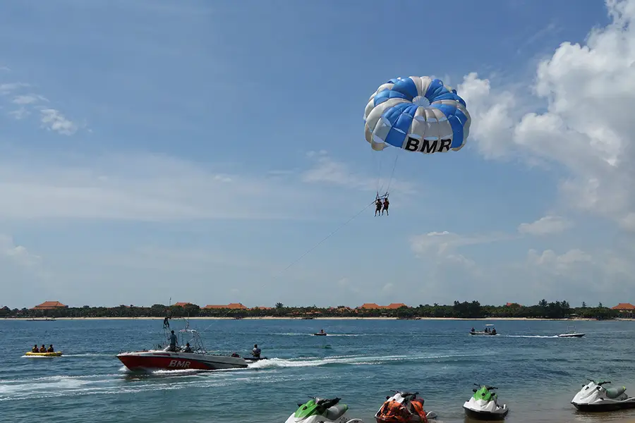 Two people parasailing high in the sky above a speedboat near a Nusa Dua resort beach.