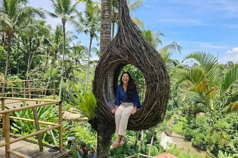 A smiling woman sitting inside a giant woven bird's nest structure attached to a palm tree.