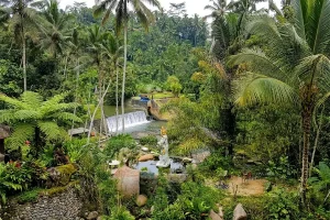 An elevated landscape view of a tropical river valley with a waterfall dam, palm trees, and a white statue.