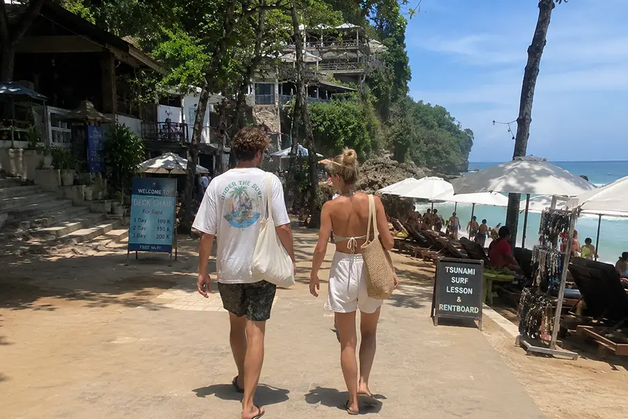 A couple walking along a beachside pathway lined with surfboards and cafes, wearing relaxed and simple travel outfits in bali.