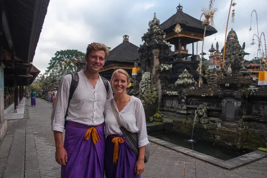 A smiling man and woman wearing traditional purple sarongs over white shirts, reflecting respectful yet simple travel outfits in bali.