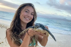 A smiling young woman gently holding a sea turtle on a sandy tropical beach.