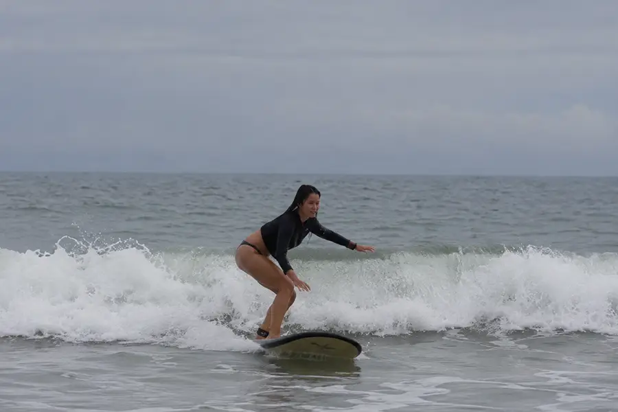 A woman in a black long-sleeve swimsuit balancing on a surfboard over a crashing ocean wave.