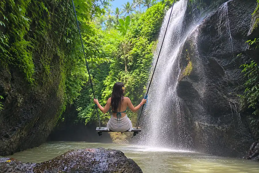 A woman sitting on a wooden swing facing a powerful waterfall inside a mossy rock canyon.