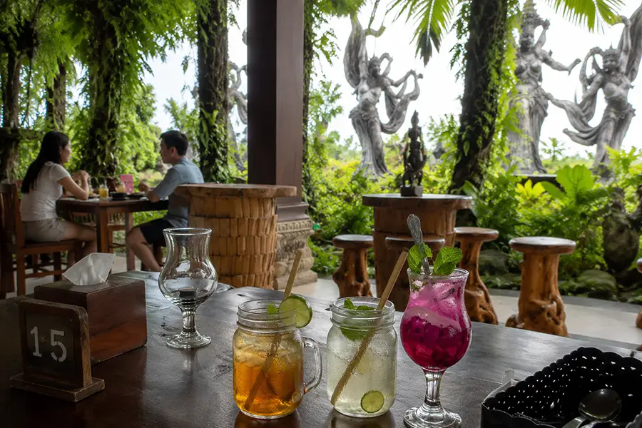 Three vibrant iced mocktails on a wooden table with diners and large stone statues visible in the background greenery.