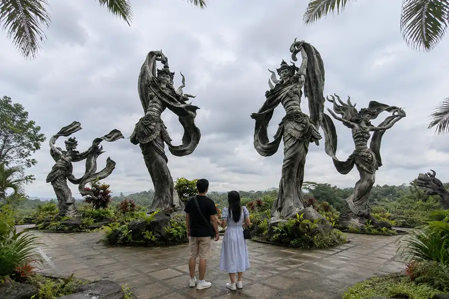 A couple from behind looking up at four giant stone angel statues standing in a landscaped garden under a cloudy sky.