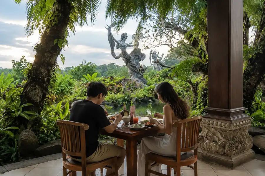 A man and a woman eating a meal at a wooden table inside an open pavilion overlooking a green pond and a giant stone statue.