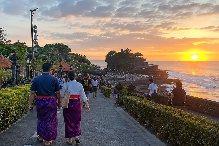People walking along a paved trail past traditional Balinese split gates and orange-roofed shrines.