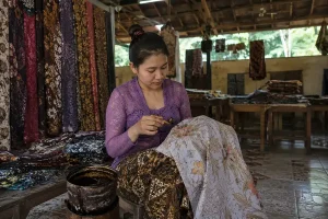 A woman in a purple lace blouse carefully applying hot wax onto a white fabric sheet to create a floral batik pattern.