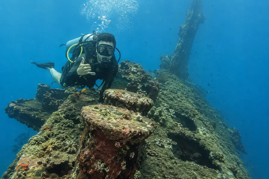 A scuba diver giving a shaka hand sign while swimming over a coral-covered shipwreck structure.