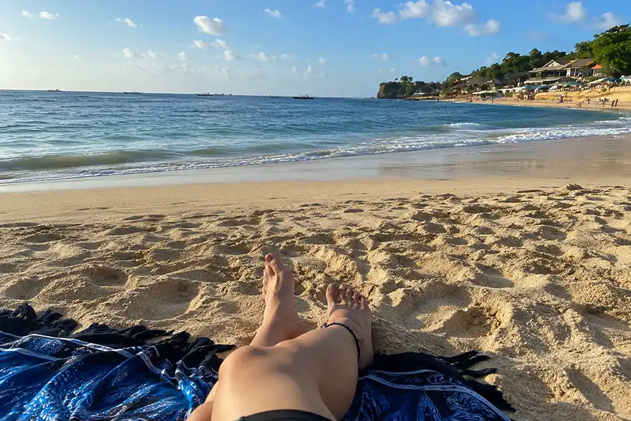 First-person view of someone relaxing on a beach towel on golden sand looking at the blue ocean.