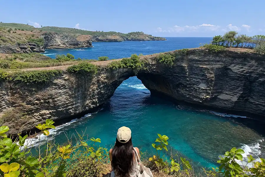 A woman sitting on a cliff looking down at the iconic archway rock formation and turquoise cove.