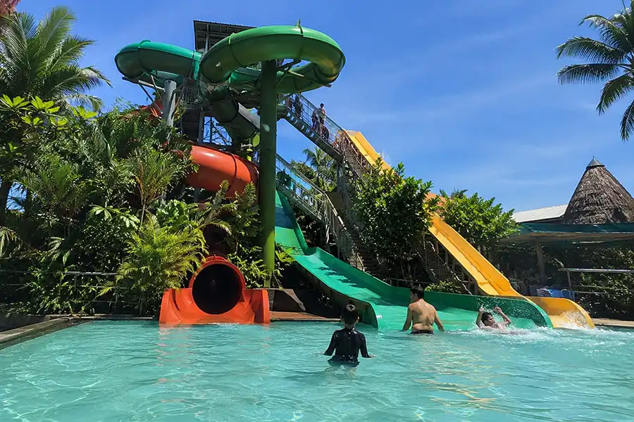 Children playing in a clear swimming pool at the bottom of multiple green, orange, and yellow water slides surrounded by tropical trees.