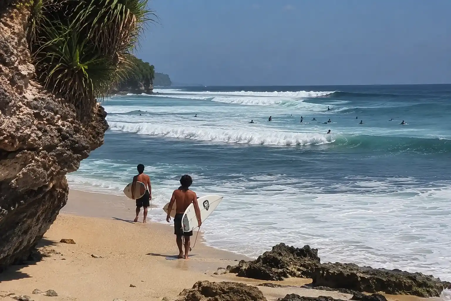 A view from a rocky cliff over a sandy beach with two shirtless surfers carrying boards towards powerful, rolling ocean waves under a blue sky.