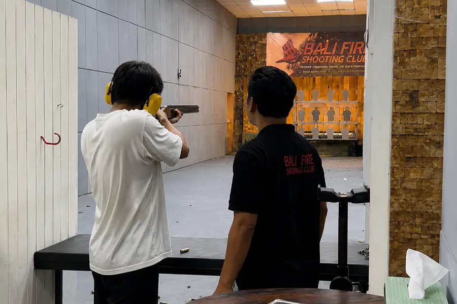 A man aiming a rifle at target sheets in an indoor range accompanied by an instructor.