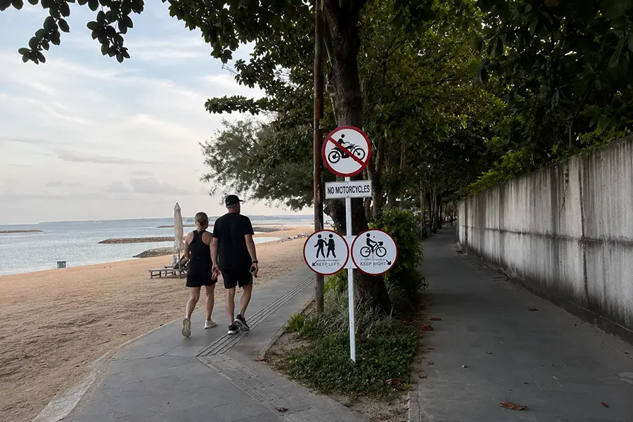 A couple walking along a paved beachfront path next to a sandy shore and clear pedestrian directional signs.