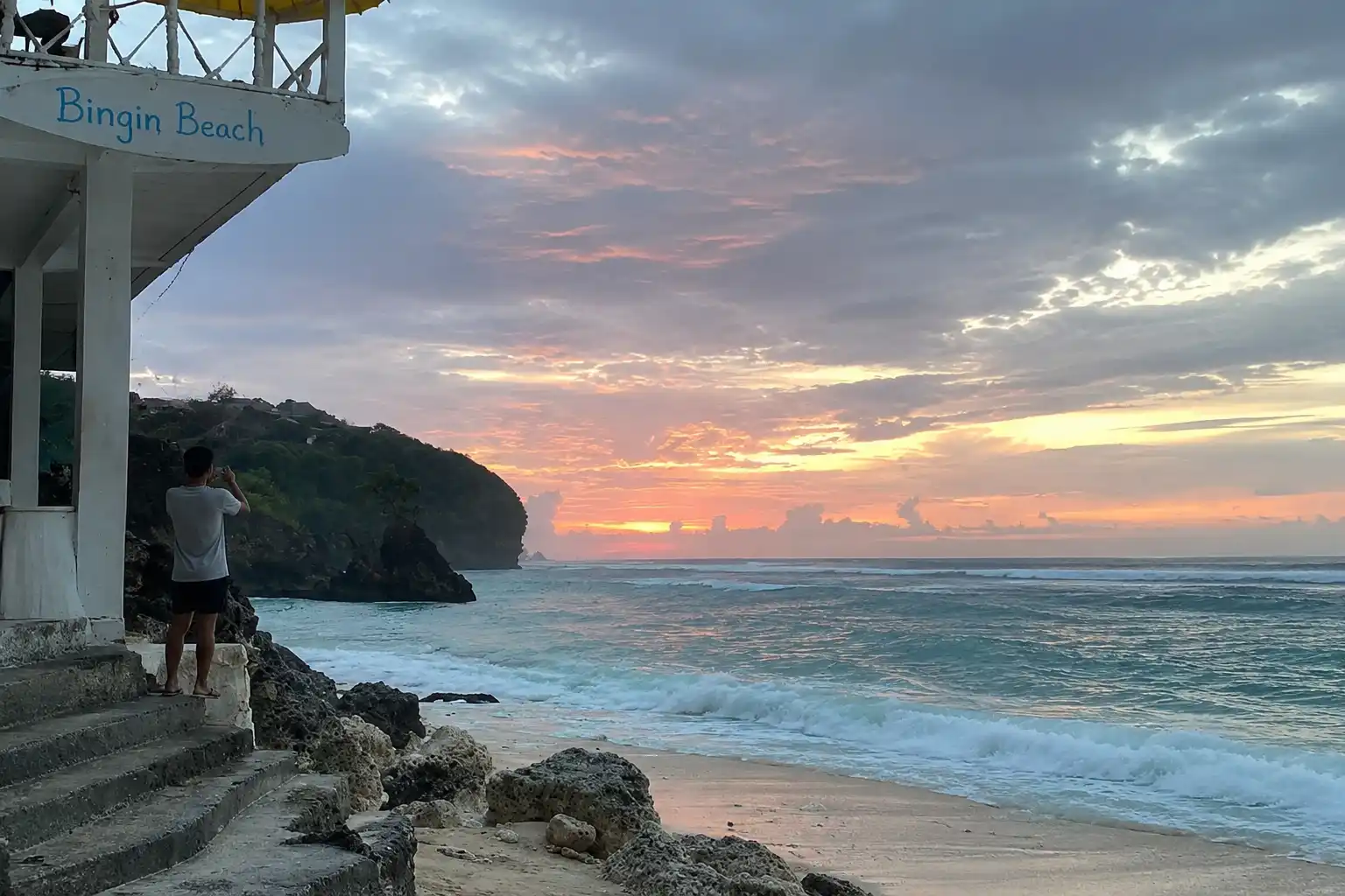 A person taking a photo of a golden sunset over ocean waves from a cliffside balcony at Bingin Beach.